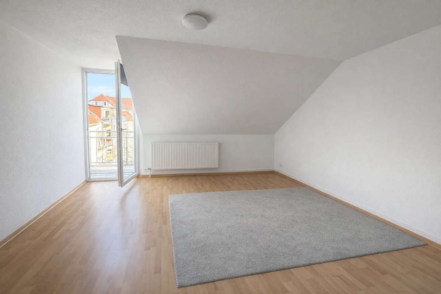 Bright attic room with white walls, wood floor, and gray rug. Balcony door open to a view of buildings.