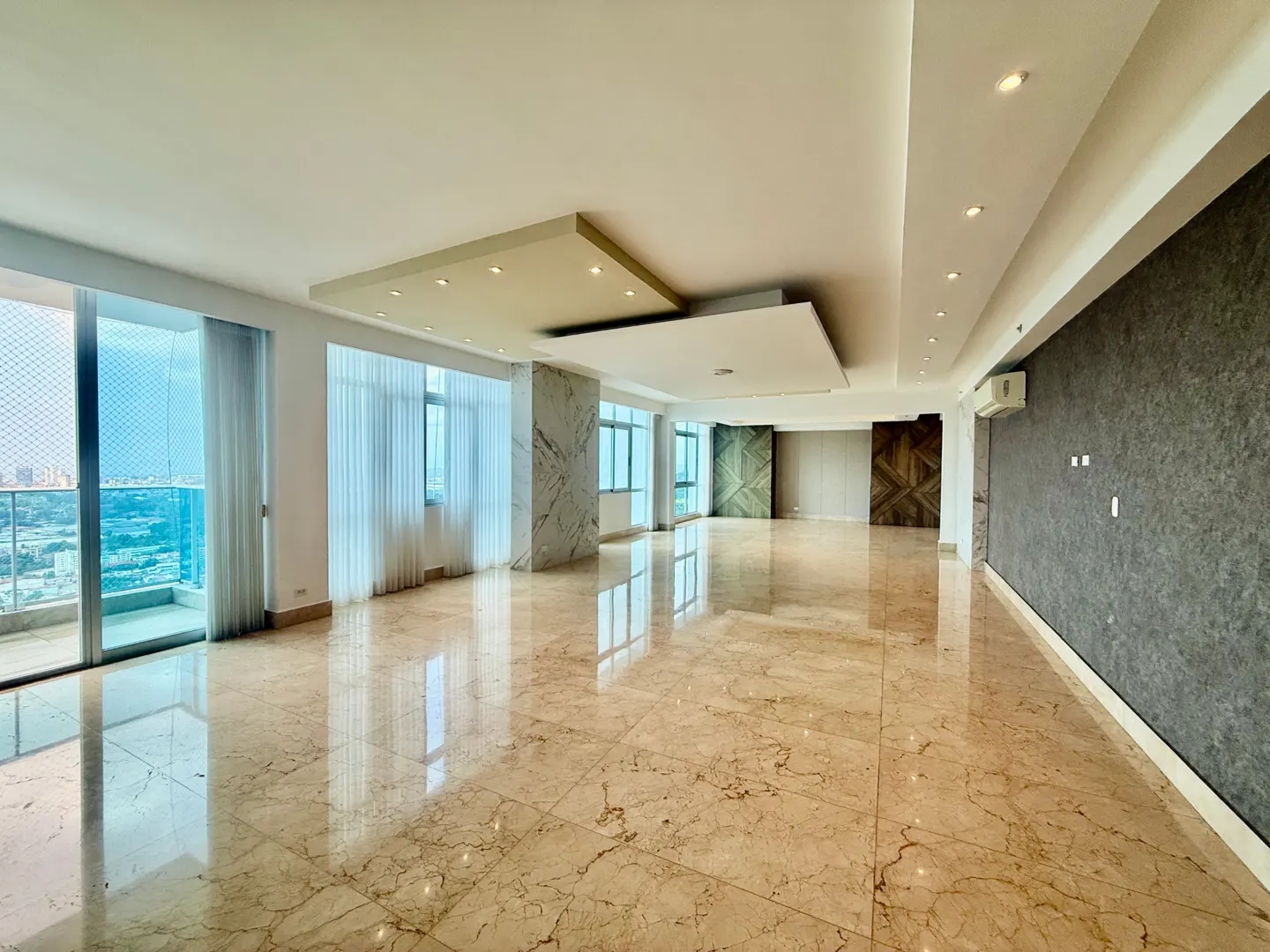 A spacious, empty apartment with marble floors, large windows with sheer curtains, and a textured gray wall.