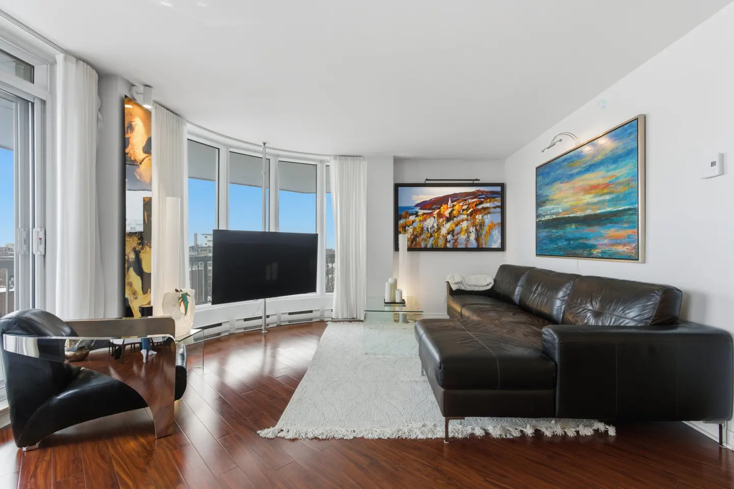 Bright living room with wood floors, a black leather sectional, and a large curved window with white curtains. Two paintings hang on the wall.