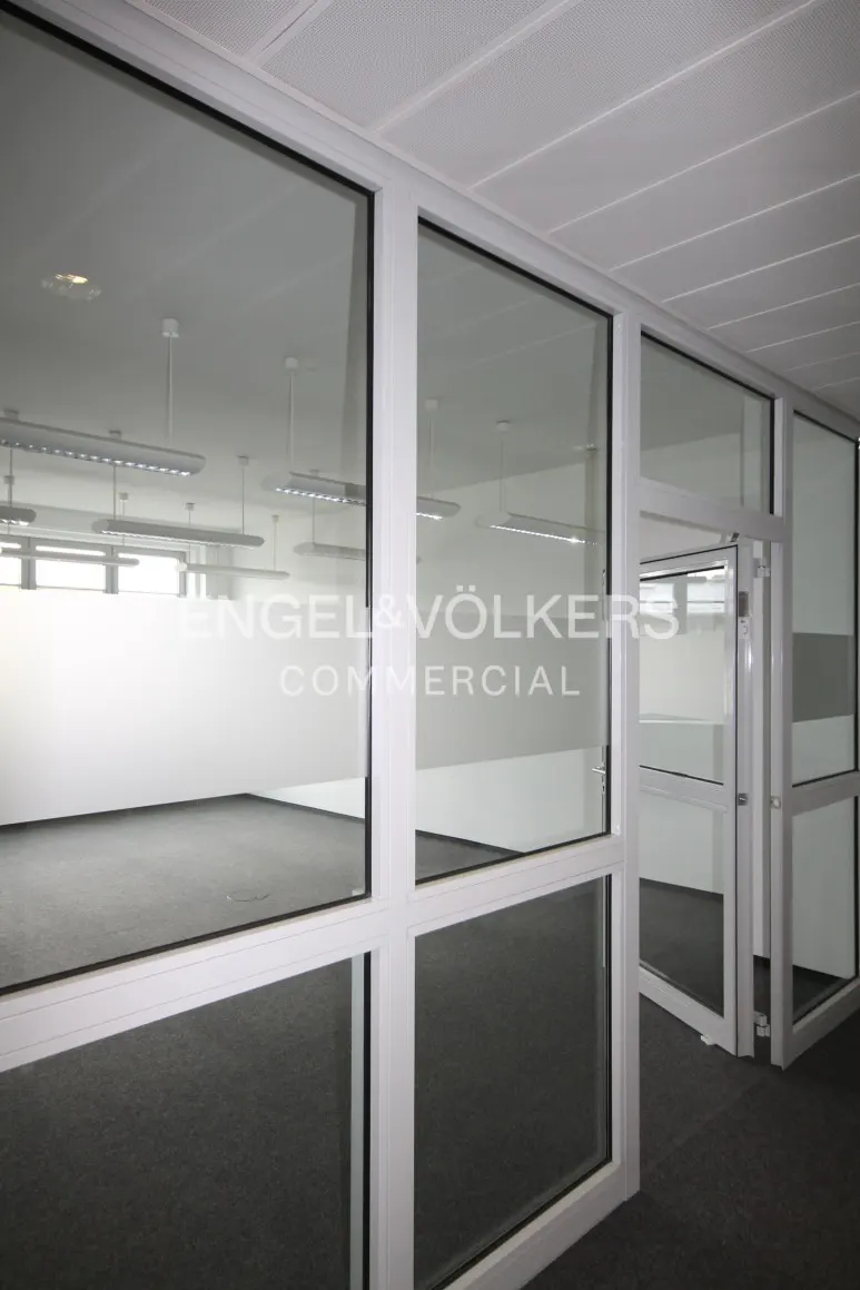 Interior view of a commercial space with white walls, gray carpet, and glass-paneled walls with white frames. "Engel & Völkers Commercial" logo on glass.