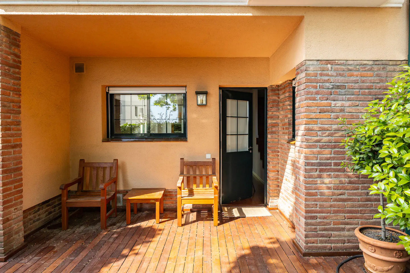 Outdoor patio with two wooden chairs, a small table, and an open black door. Brick and stucco walls, a window, and a potted plant are visible.