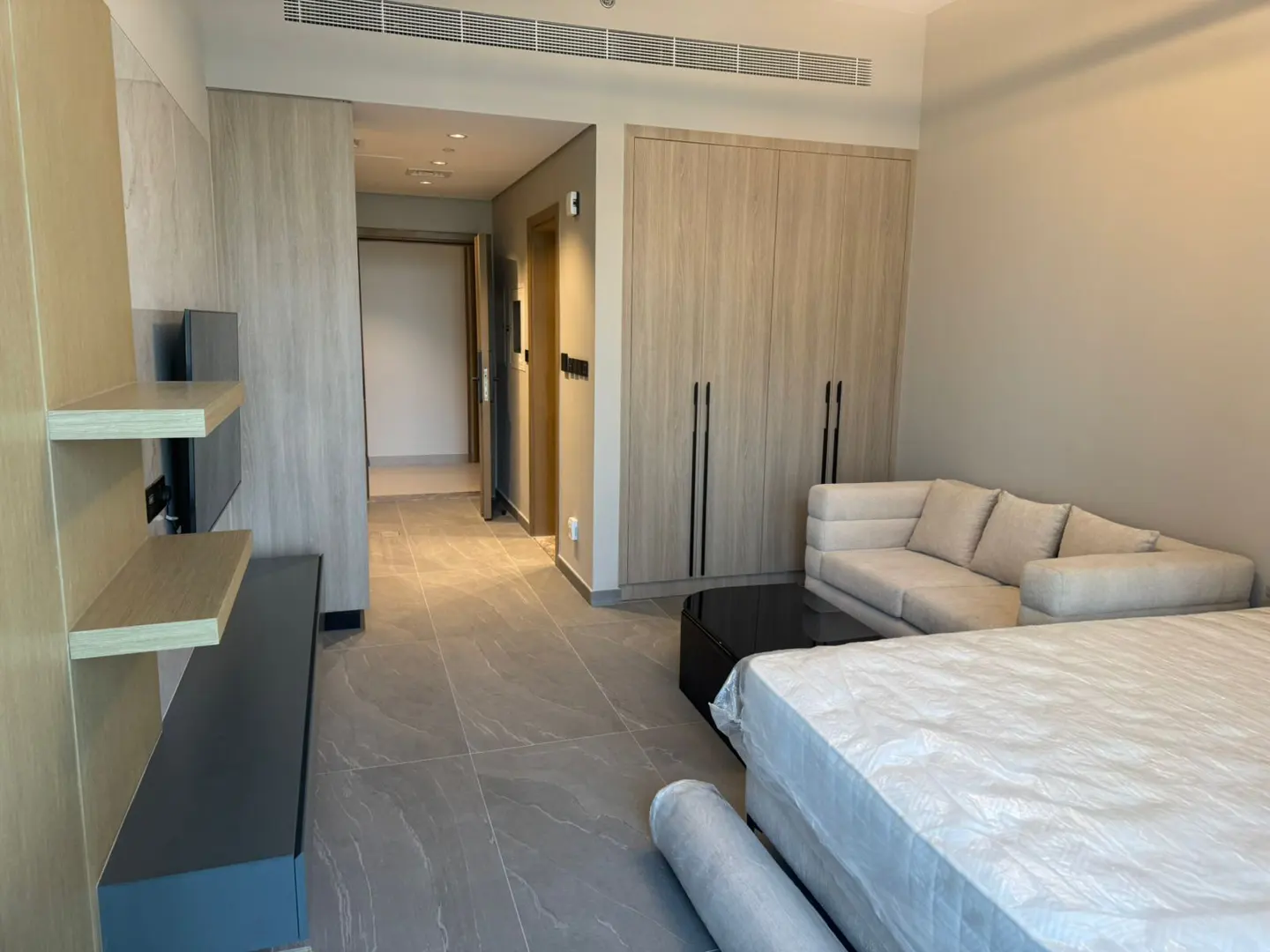 A modern apartment interior with light wood cabinets, a beige sofa, and gray tile flooring. A bed is wrapped in plastic.