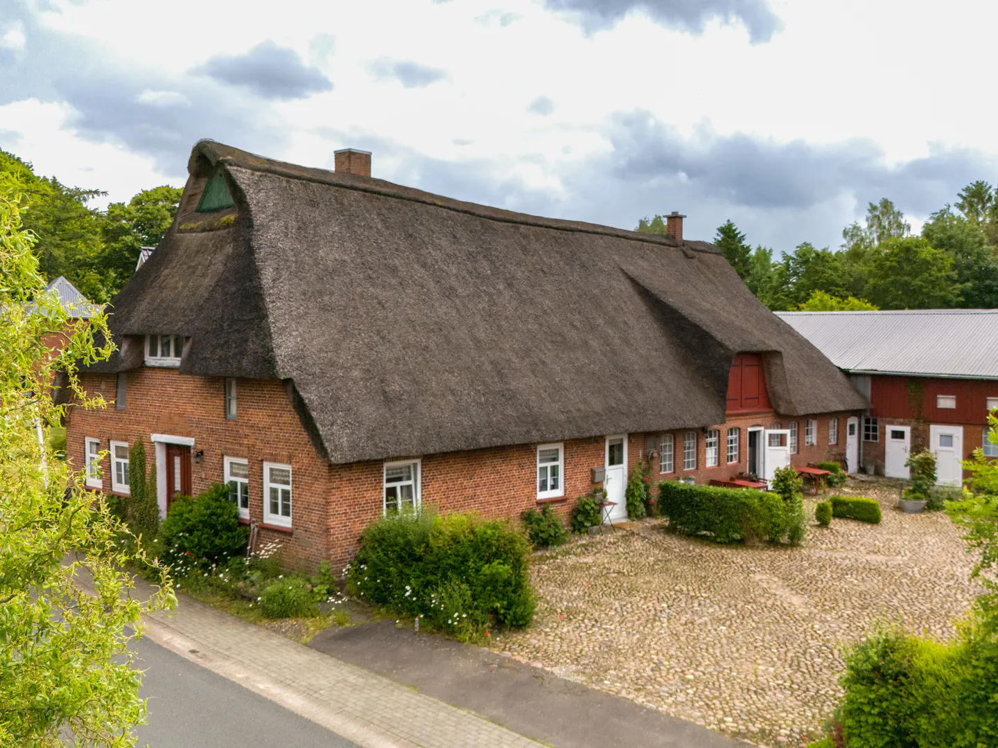 Exterior view of a red brick house with a thatched roof, white windows, and a cobblestone courtyard.
