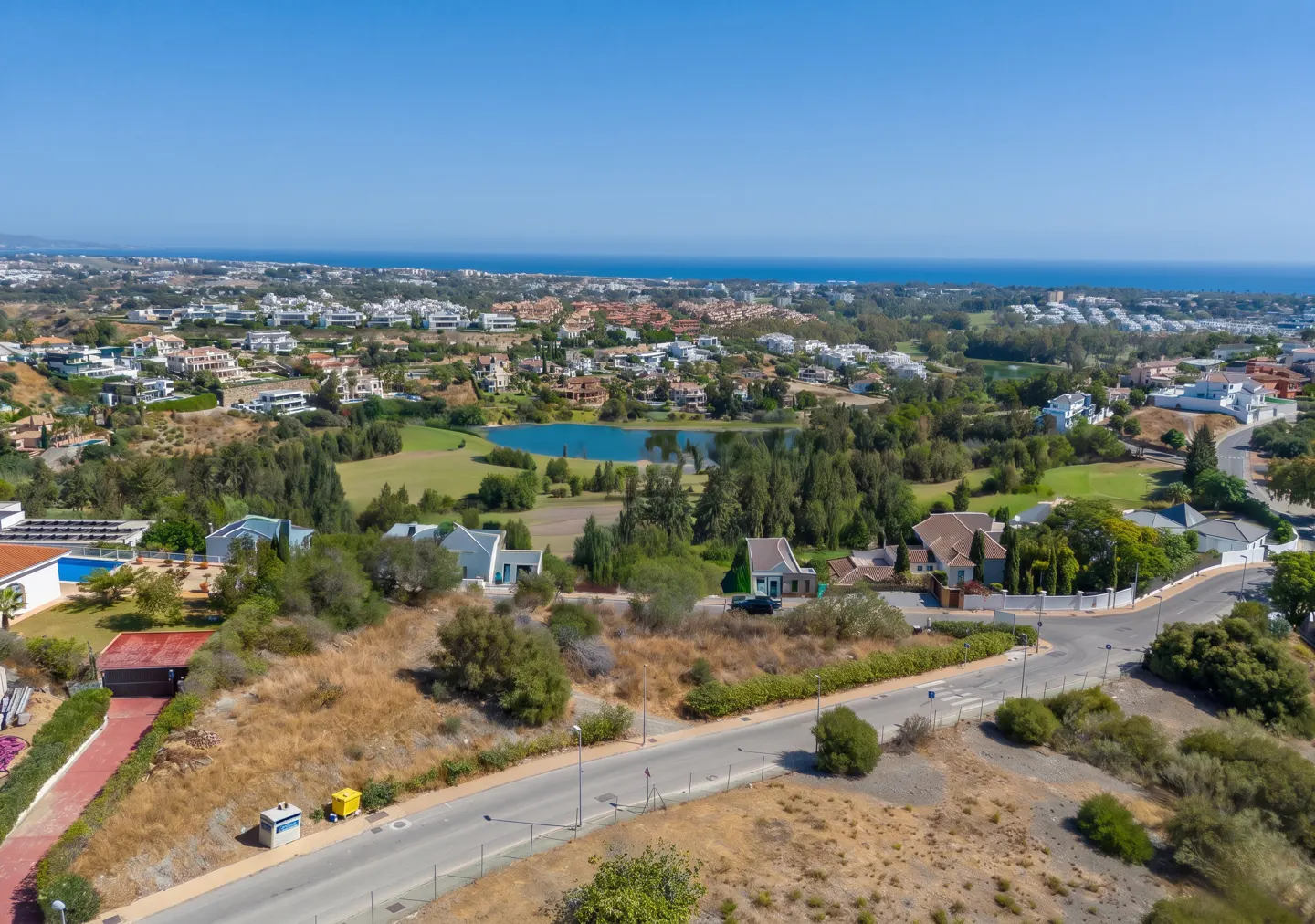 Aerial view of a residential area with houses, trees, a golf course with a pond, and the ocean in the background under a clear blue sky.