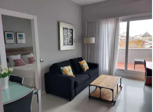 Living room with a black sofa, wooden coffee table, and balcony with city views. Bedroom visible through a doorway.
