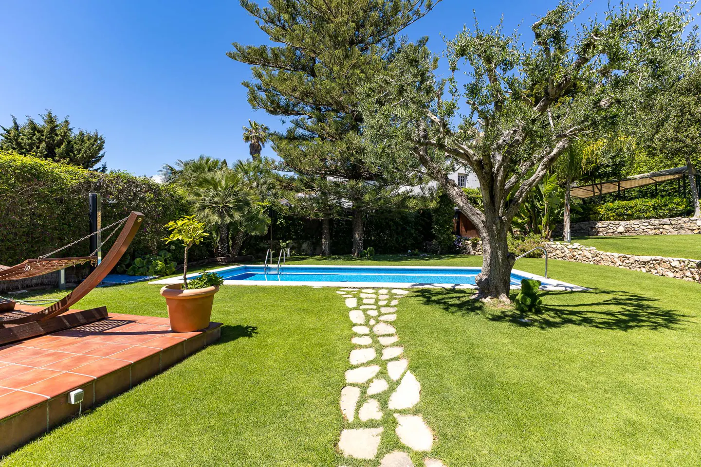 A backyard with a pool, hammock, stone path, and green grass under a blue sky.