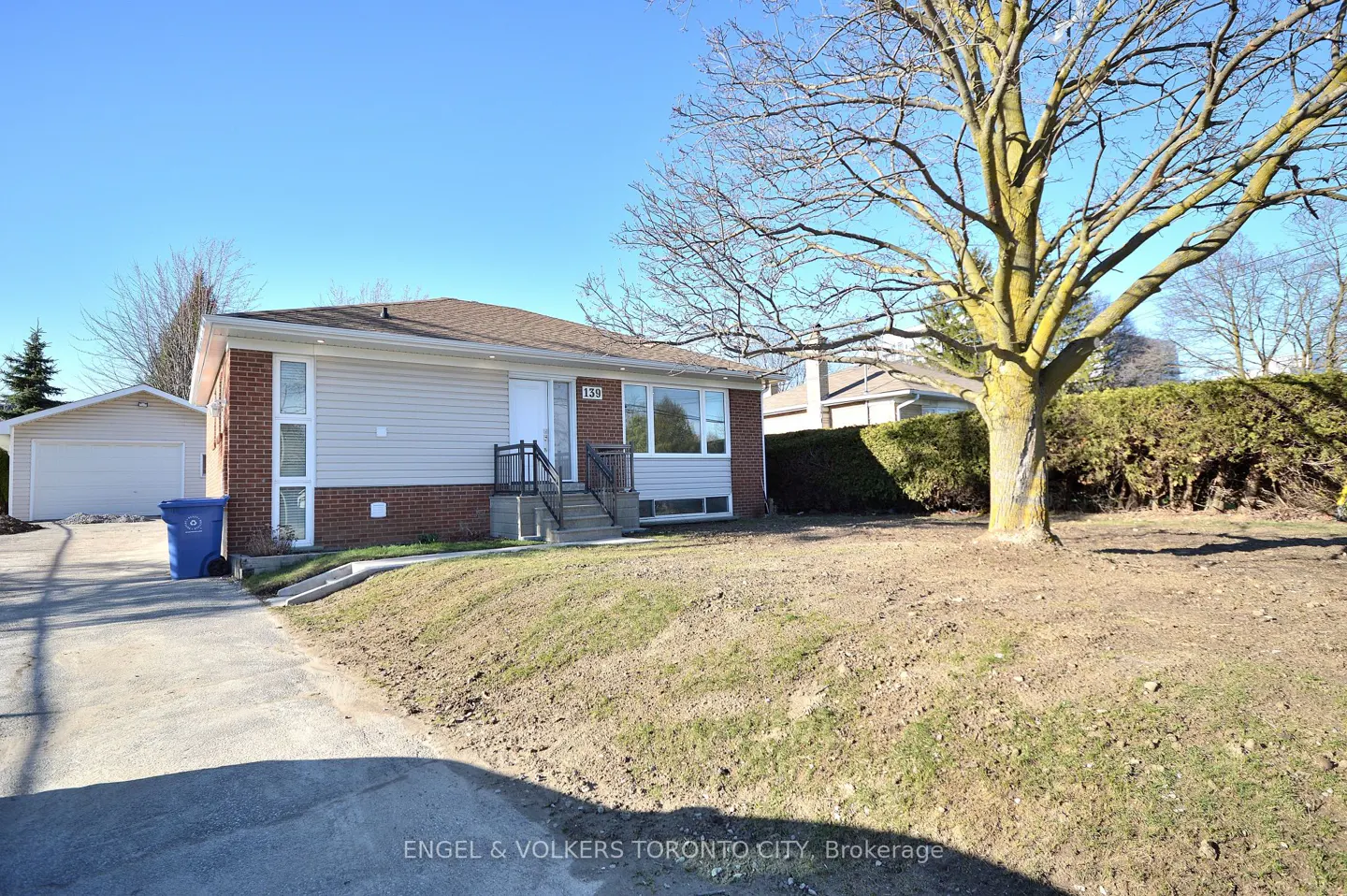 Exterior view of a one-story house with beige siding, brick accents, and a brown roof on a sunny day. A large tree stands in the front yard.