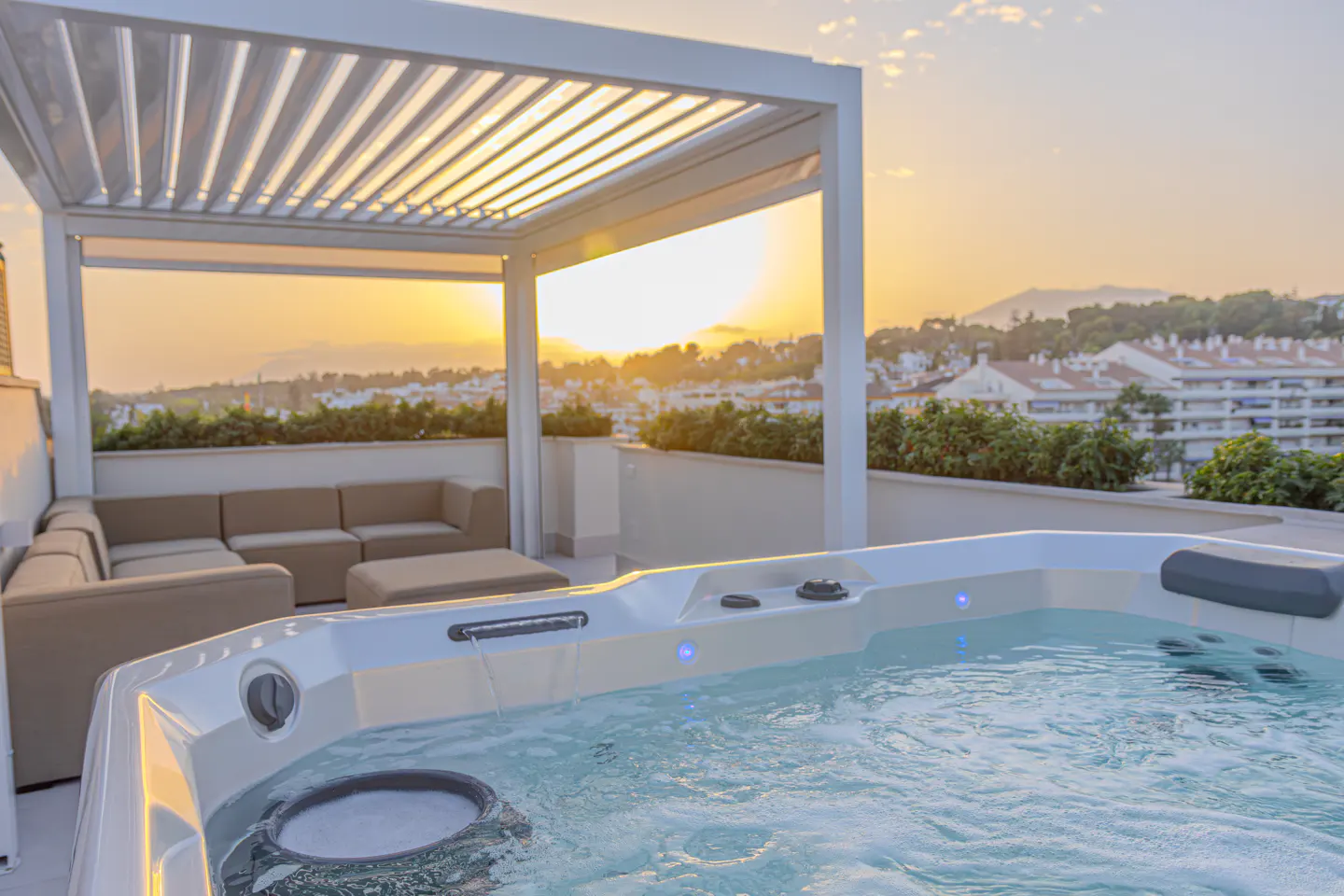 Rooftop terrace with a white pergola, tan sectional sofa, and a bubbling hot tub overlooking a city at sunset.
