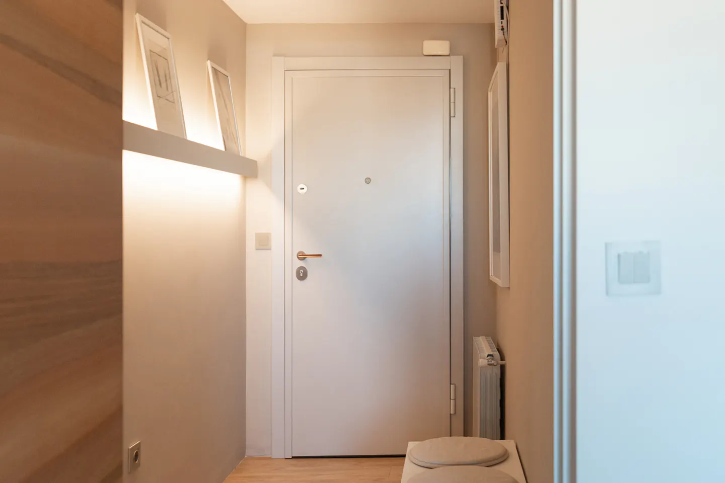 Entryway with a white door, gold handle, and peephole. A shelf with framed art is illuminated by a light strip. A radiator and bench are near the door.