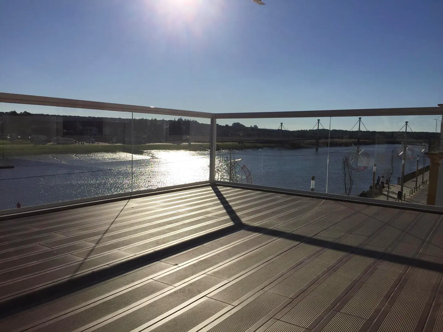 A wooden deck with a glass railing overlooking a sunlit river and a distant bridge under a clear blue sky.