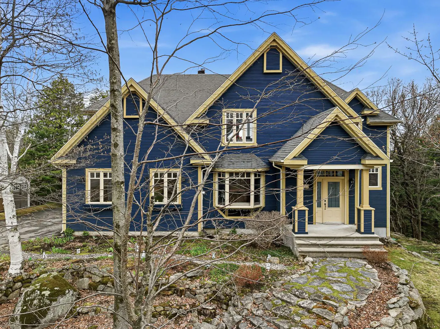 A two-story blue house with yellow trim, a gray roof, and a light yellow front door. The house is surrounded by trees and rocks.