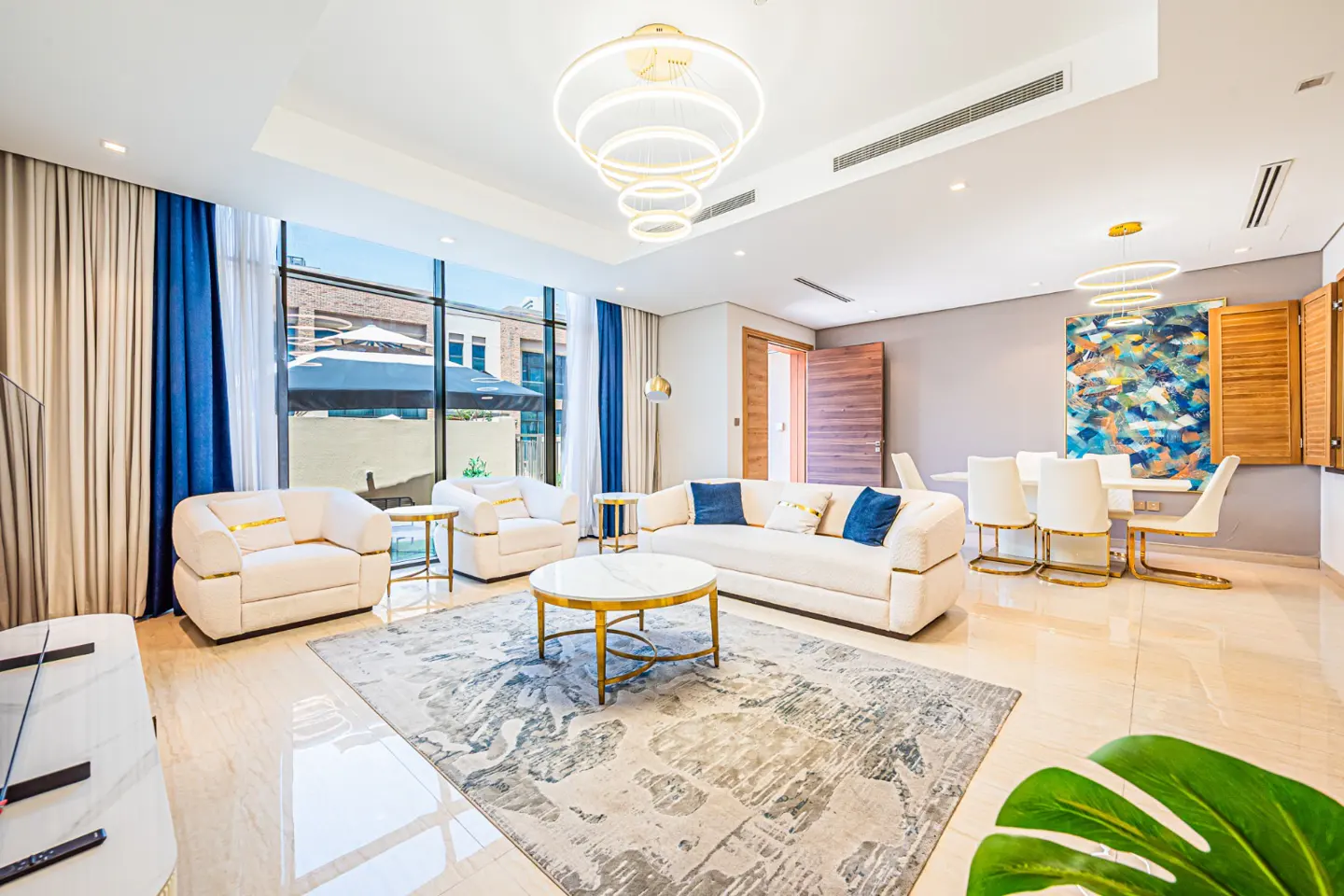 Bright, modern living room with white sofas, a marble table, and a large window with blue curtains. Dining area with white chairs is visible.