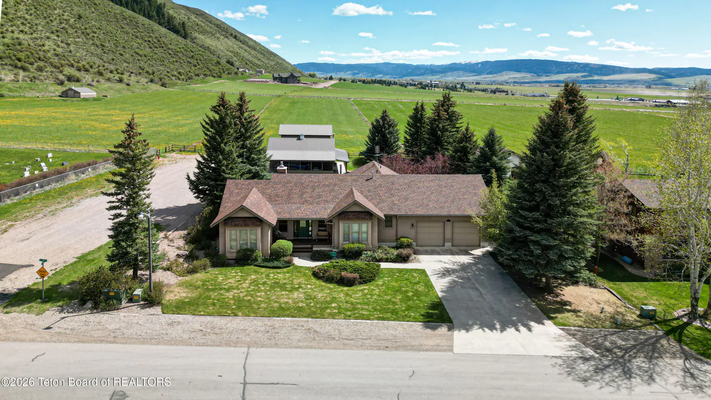 Tan house with a brown roof, green lawn, and driveway. Trees surround the house, with a mountain and field in the background.
