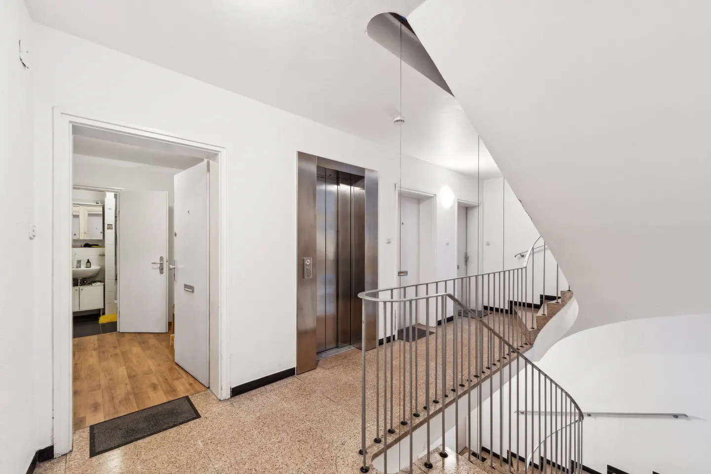 Interior view of a building hallway with an elevator, stairs with metal railings, and white walls.