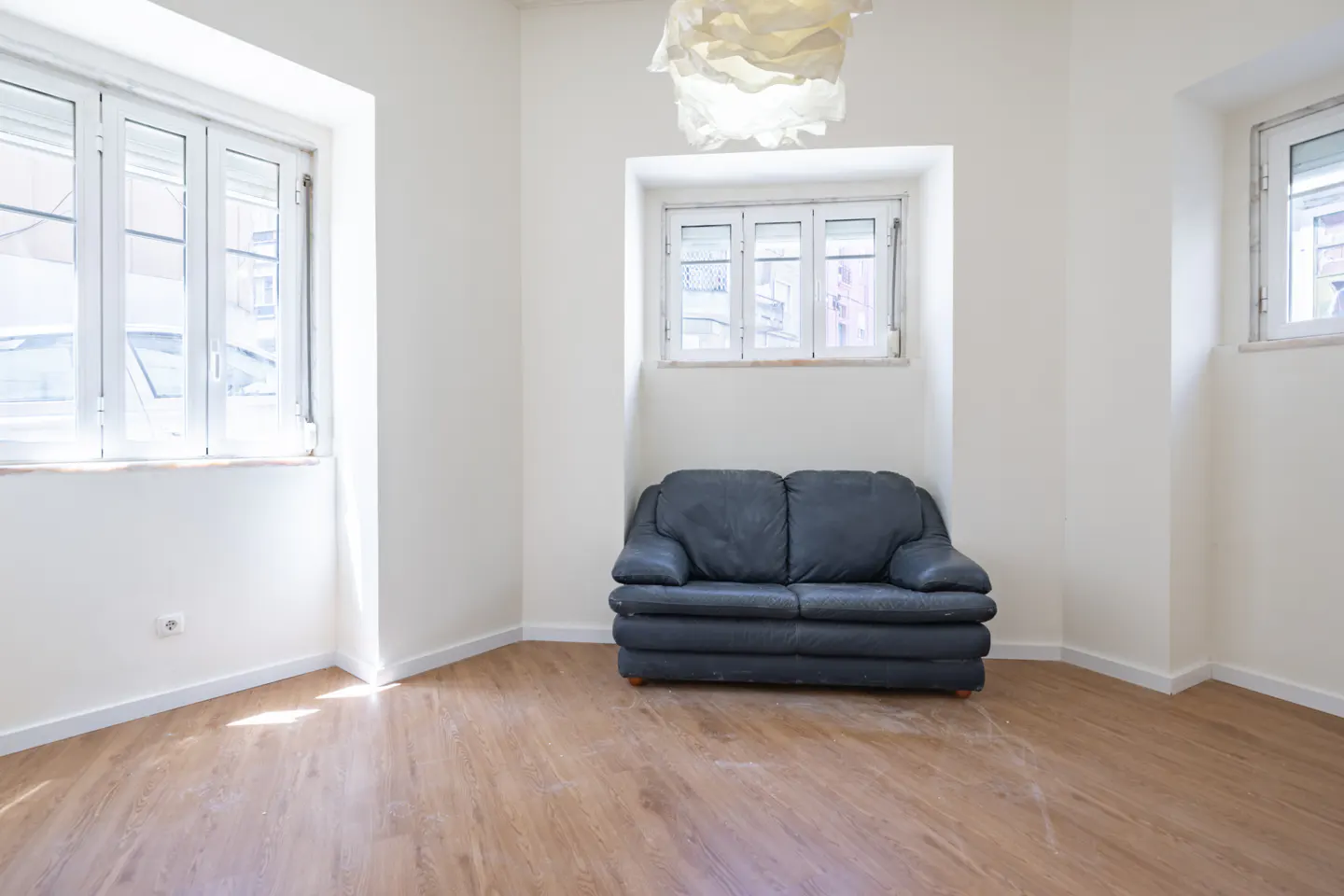 Bright, empty room with wood floors, white walls, and three windows. A gray loveseat sits below a window with a modern white light fixture above.
