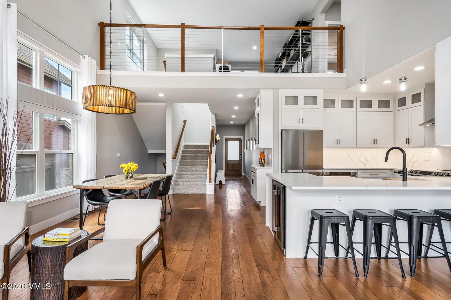 Open-concept home interior with hardwood floors, white kitchen cabinets, dining table, and a loft area above.