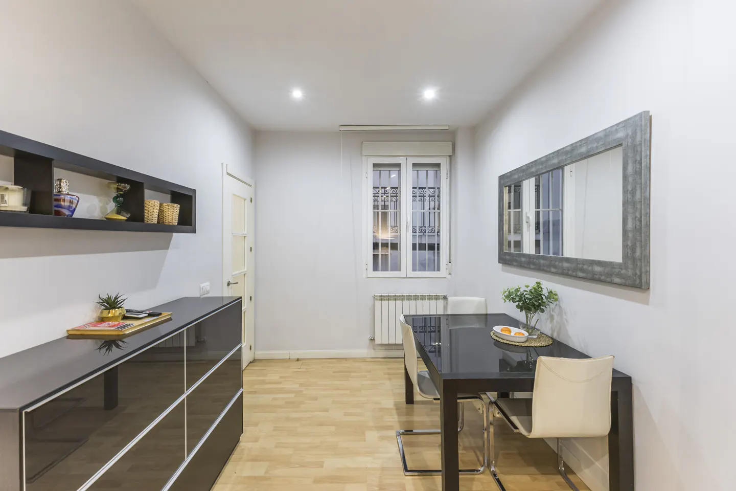 Bright dining room with a black table, white chairs, and a large mirror on the wall. A black cabinet and shelf are on the left.
