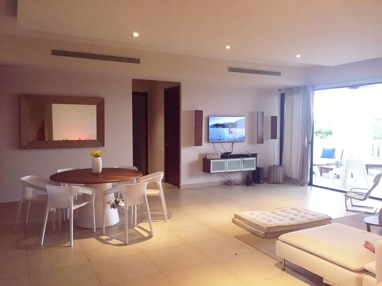 Bright, open living room with a dining table, TV, and sliding glass doors to a patio. Neutral color scheme with white furniture.