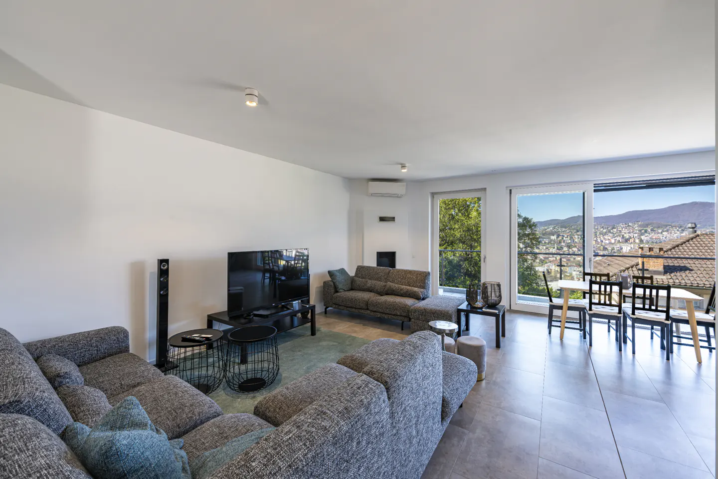 Bright living room with gray sofas, black tables, and a TV. Large windows offer a city view.