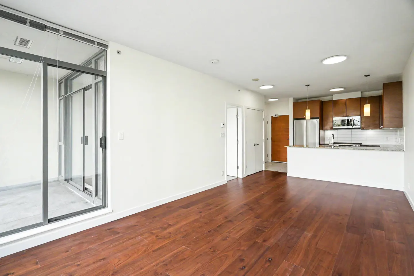 Bright, modern apartment with hardwood floors, white walls, and a kitchen with brown cabinets. Sliding glass doors lead to a balcony.