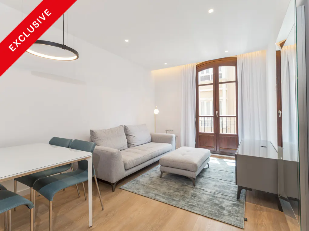 Bright living room with white walls, wood floors, and a gray sofa. A white table with blue chairs sits near a round light fixture. "Exclusive" banner in the corner.