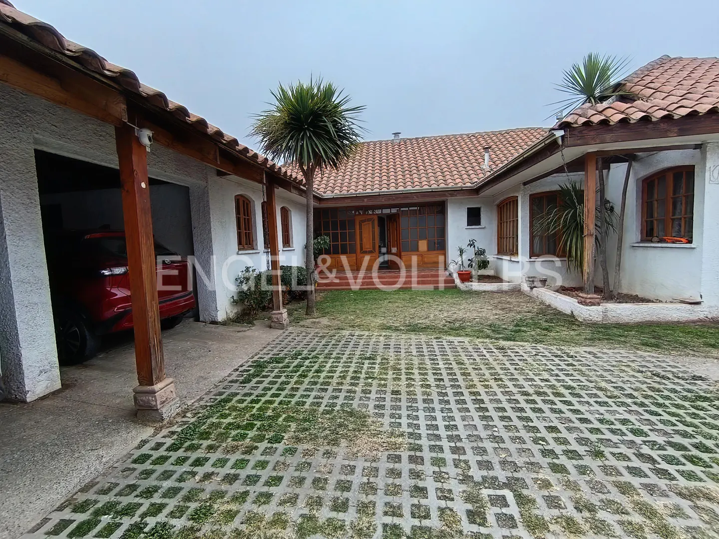 A white house with a terracotta tile roof, a covered carport with a red car, and a paved courtyard with a palm tree.