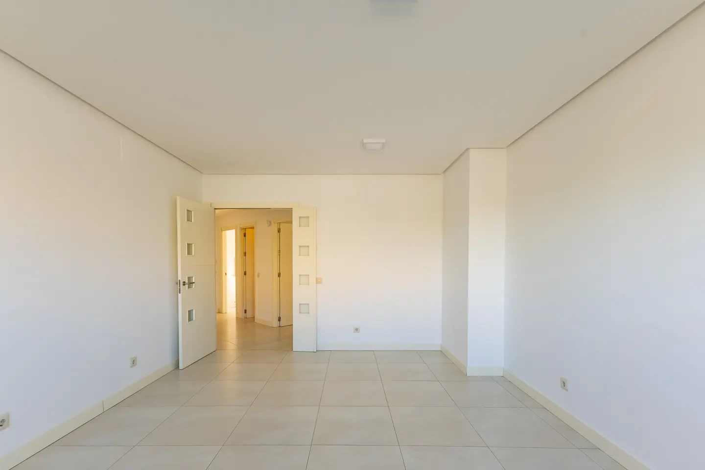 Bright, empty room with white walls and tile floor. A doorway leads to other rooms.