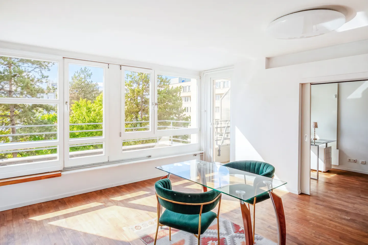 Bright dining room with wood floors, white walls, and large windows. A glass table with two green velvet chairs sits on a patterned rug.