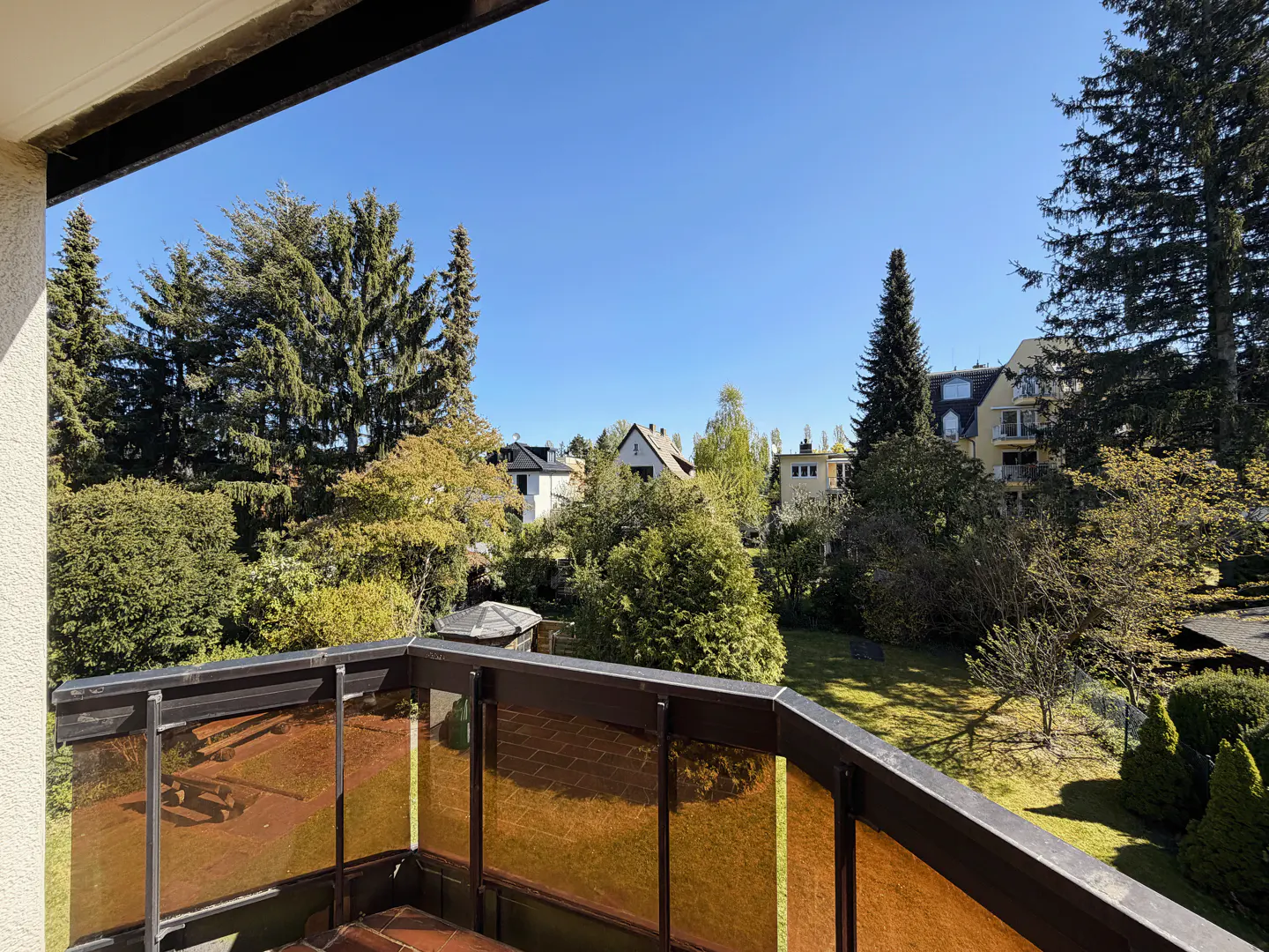View from a balcony with amber glass railings overlooking a green yard with trees and houses under a blue sky.