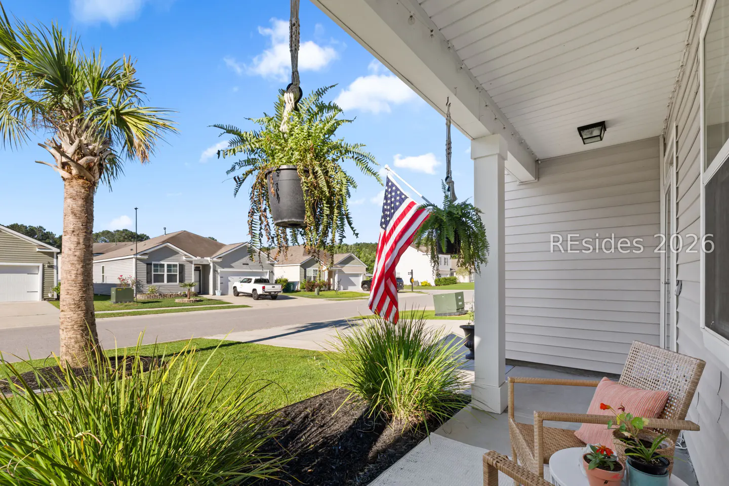 Porch view with hanging ferns, American flag, and wicker chair with a pink pillow. Palm tree and houses in the background.