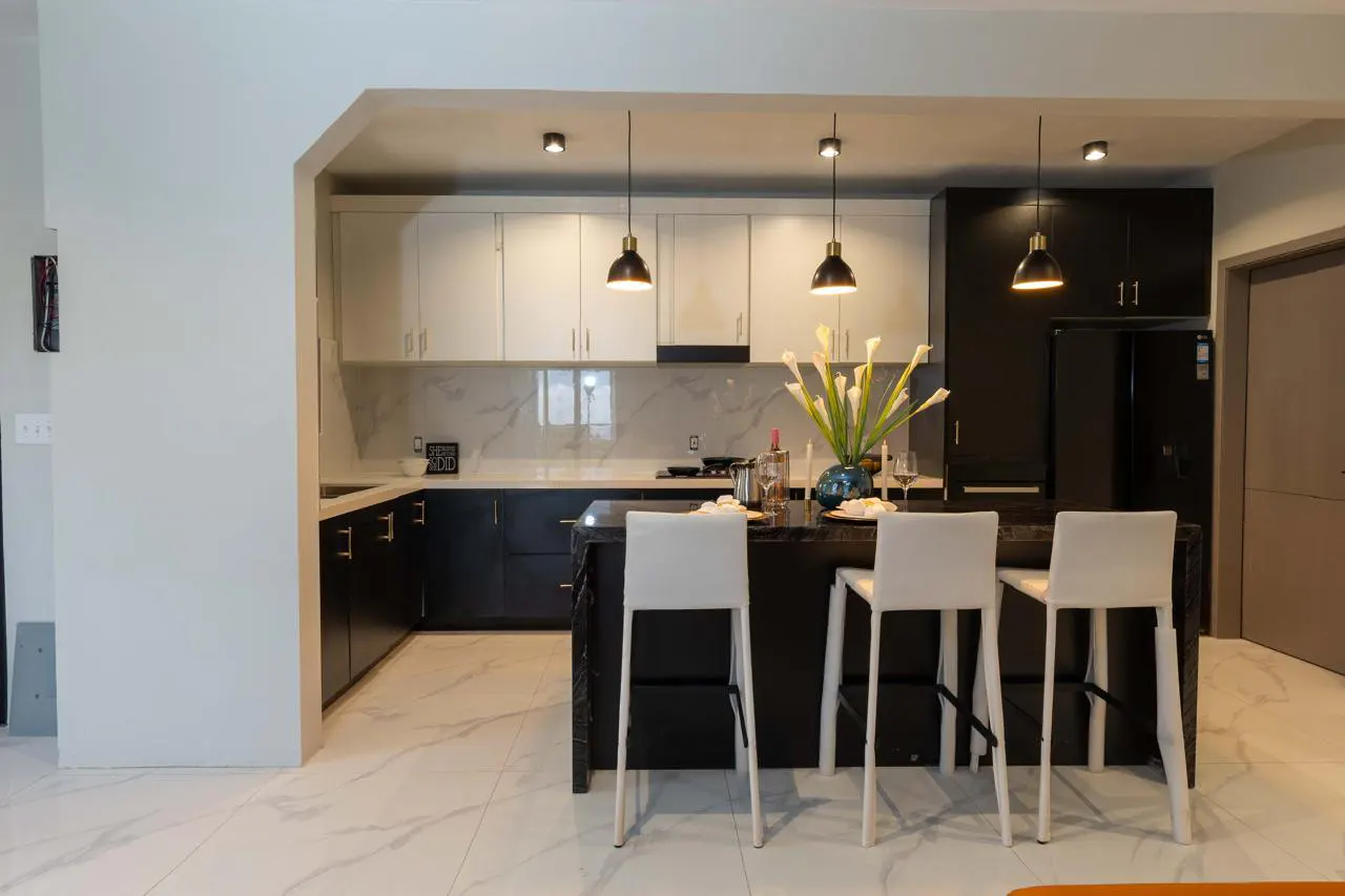 A modern kitchen with white marble floors, black island with three white stools, and black and white cabinets.