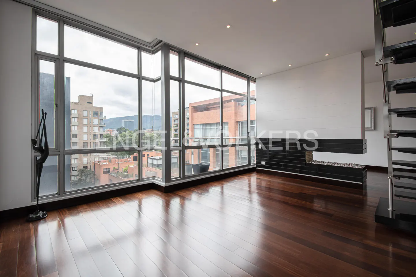 Bright, empty living room with hardwood floors, large windows showing city views, a modern fireplace, and a metal staircase.