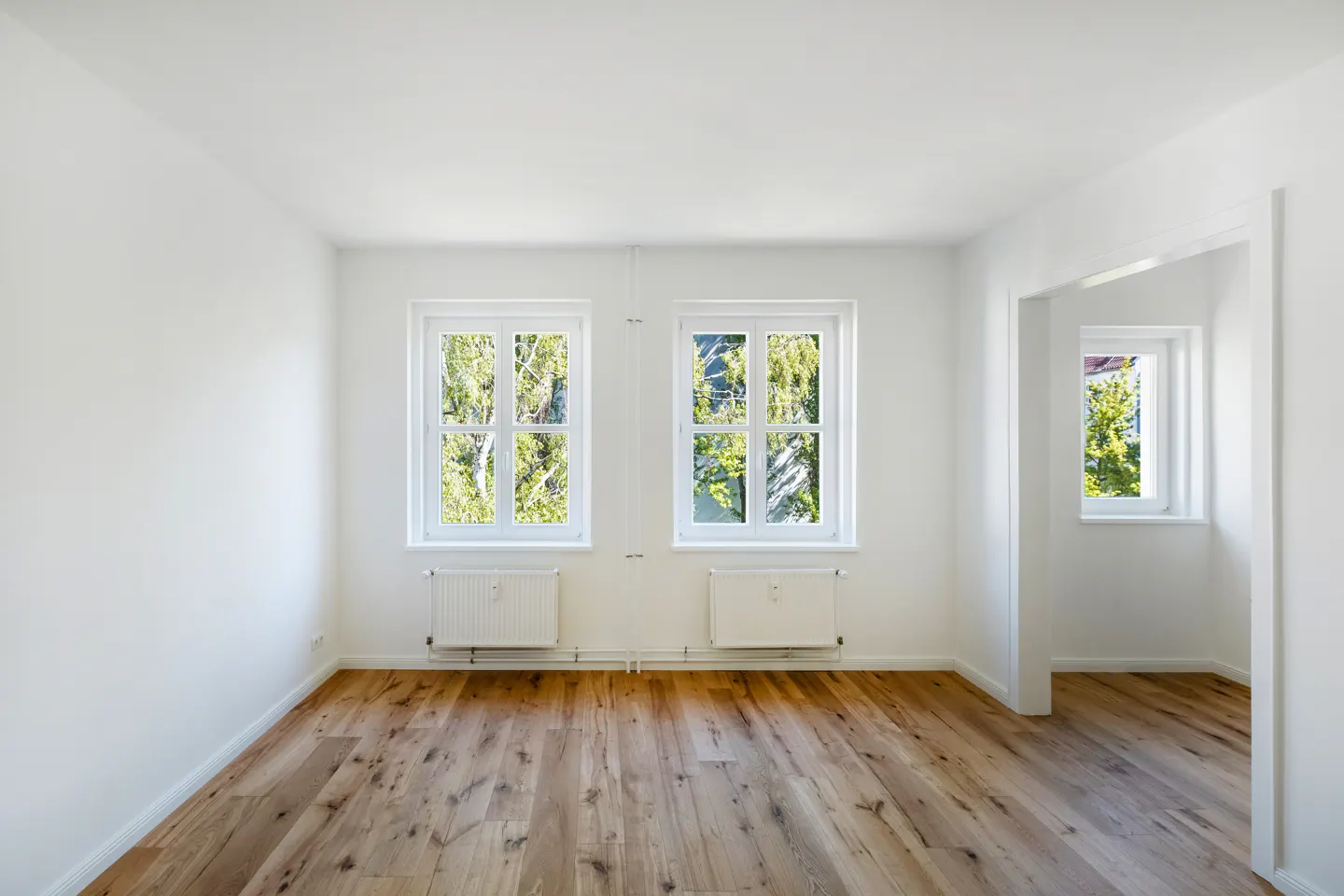 Bright, empty room with hardwood floors, white walls, two windows, and a doorway to another room.