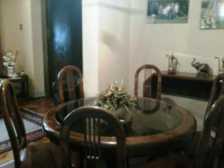 A dining room features a round, glass-topped table with wooden chairs and a floral centerpiece. A dark wooden door and wall art are in the background.