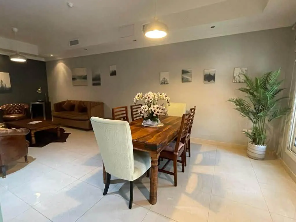 A dining room with a wooden table, chairs, and a floral centerpiece. A living room area with sofas and a coffee table is in the background.