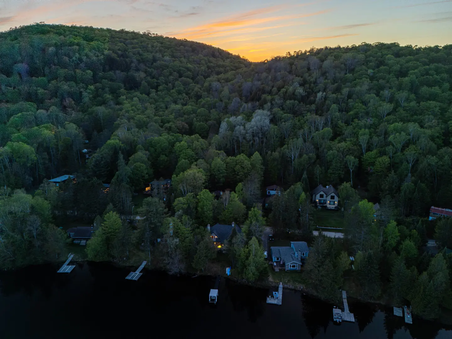 Aerial view of lakefront homes nestled in trees at dusk, with a hill in the background and a colorful sunset.