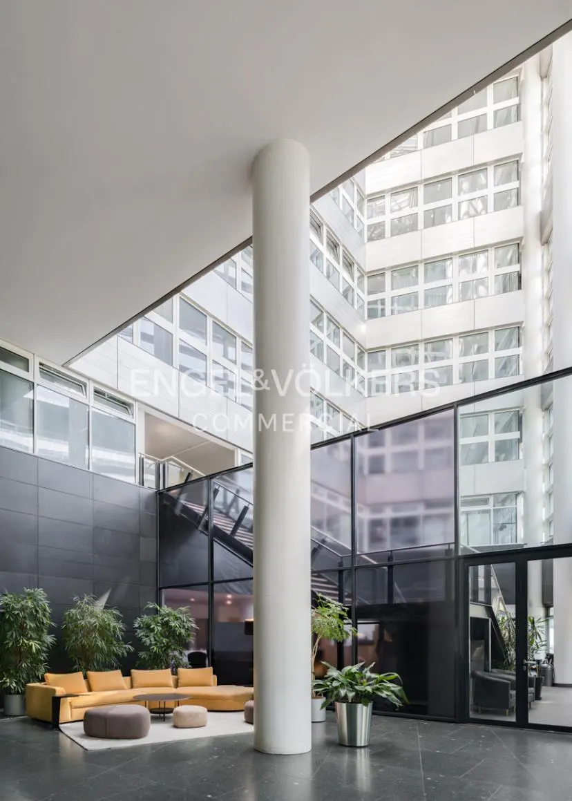 Bright lobby with a yellow sofa, plants, and a large white pillar. The building's facade is visible through the open ceiling.