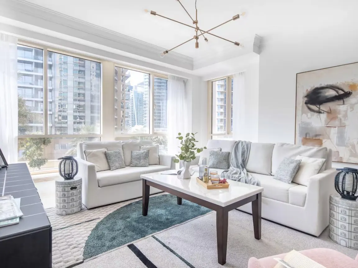 Bright living room with white sofas, marble coffee table, and large windows overlooking city buildings. Modern chandelier hangs from the ceiling.
