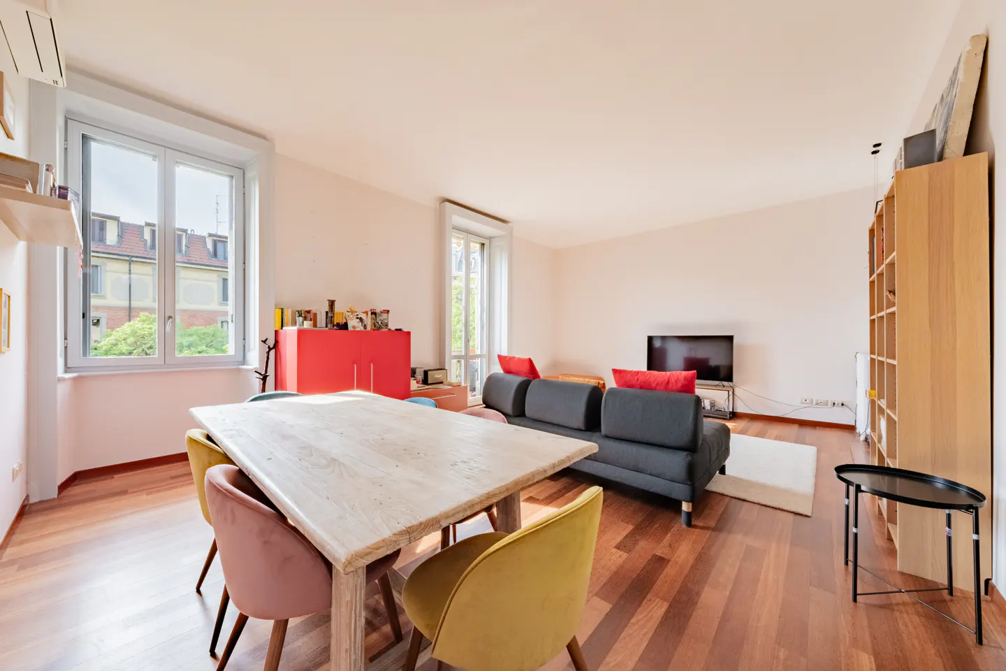 Bright living room with wood floors, a dining table with pink and yellow chairs, and a gray sofa with red pillows.