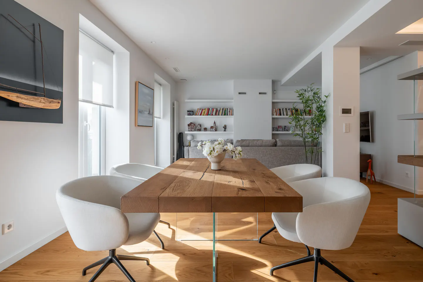 Bright dining room with a wooden table, glass legs, white chairs, and a vase of white flowers. Bookshelves line the back wall.