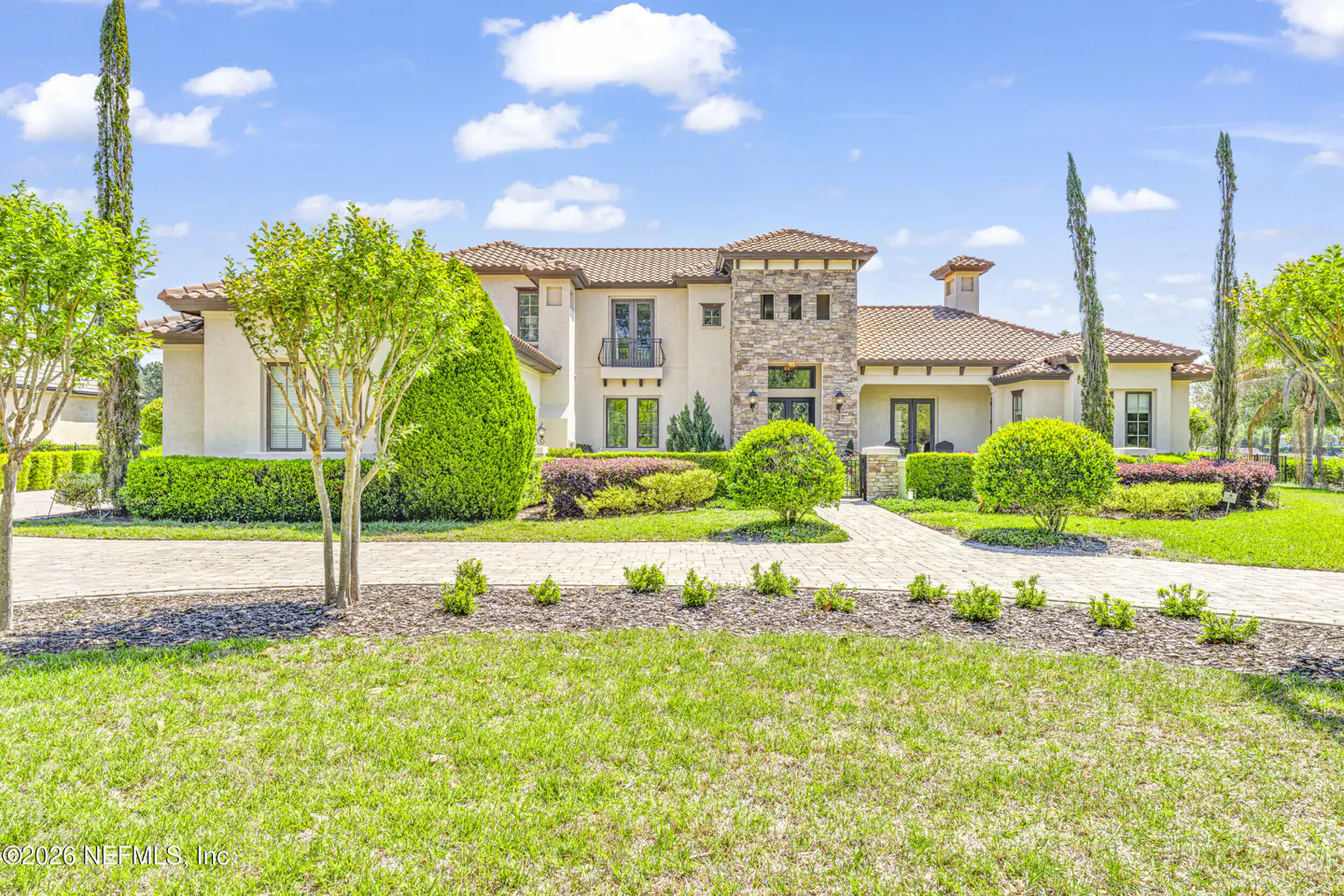 Beige two-story house with a brown tile roof, stone accents, and manicured green landscaping under a blue sky.