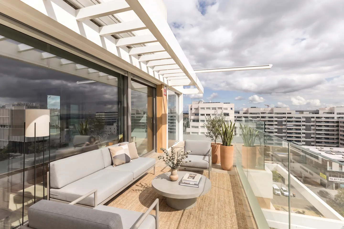 A modern balcony with gray furniture, a round coffee table, and potted plants on a jute rug, with city views.