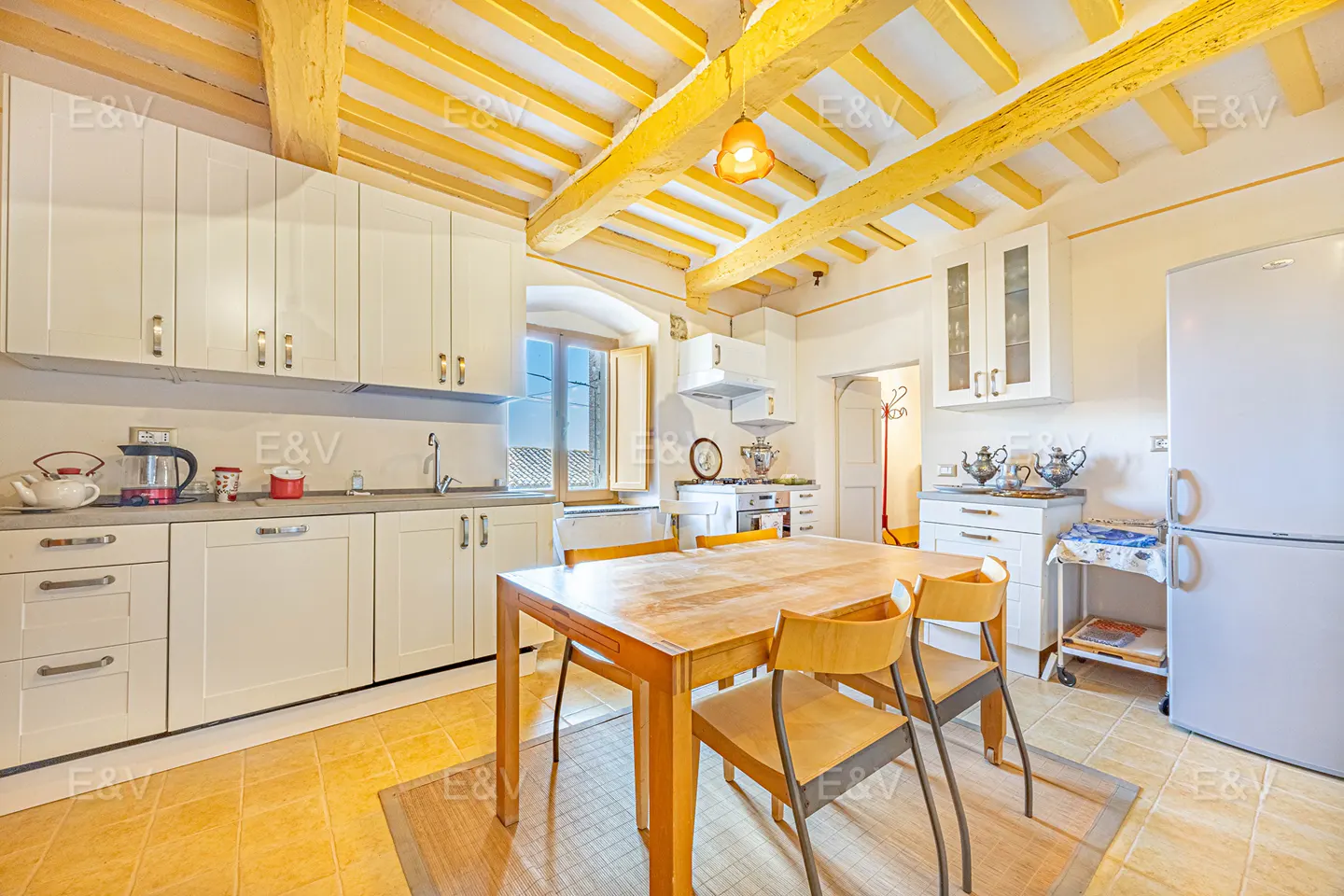 Bright kitchen with white cabinets, yellow ceiling beams, and a wooden table with four chairs. A white refrigerator stands to the right.