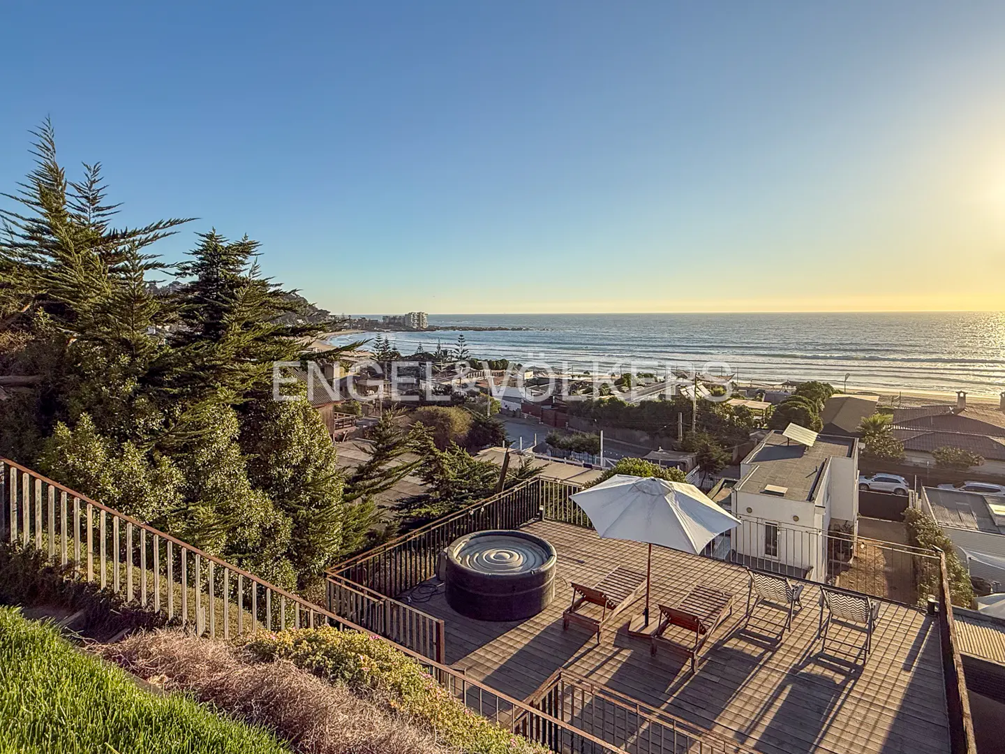 Rooftop deck with a hot tub, lounge chairs, and umbrella overlooking the ocean and beach at sunset.