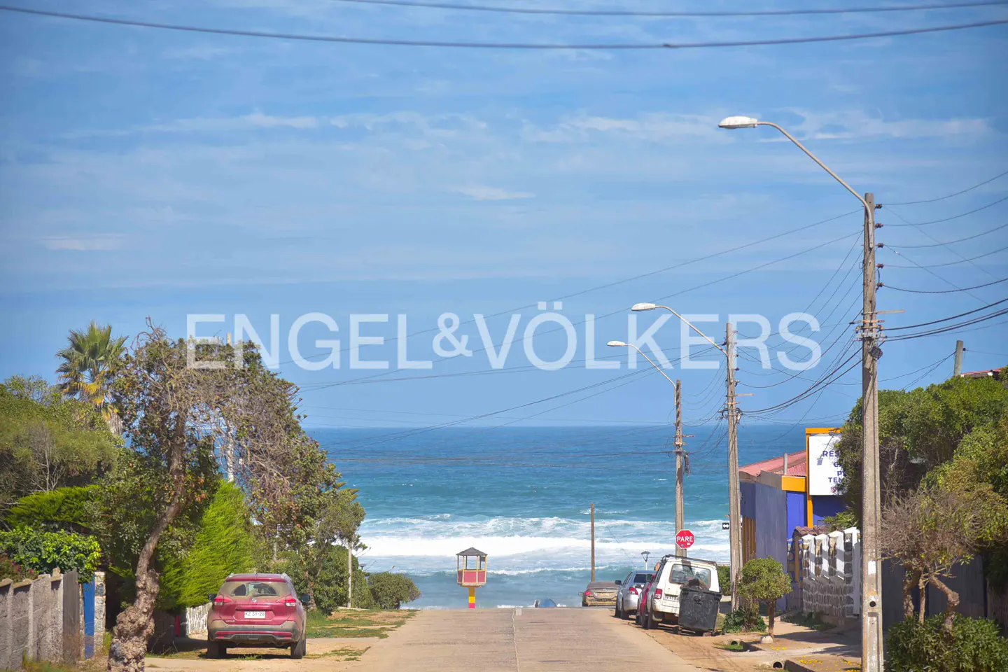 Street view leading to the ocean, with parked cars, utility poles, and lush greenery on either side. Blue sky above.