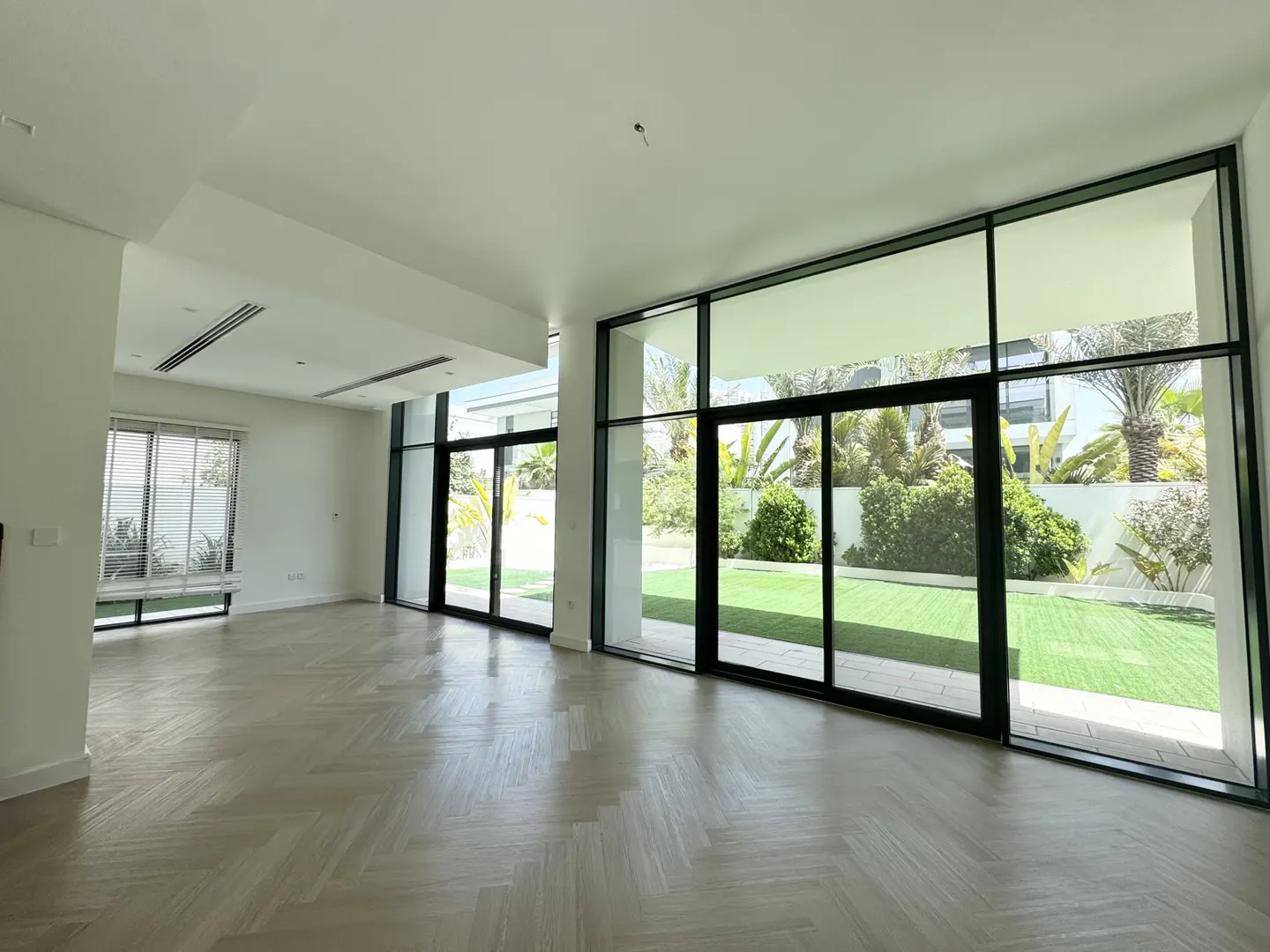 Bright, empty living room with herringbone wood floors, white walls, and large black-framed windows overlooking a green lawn and garden.