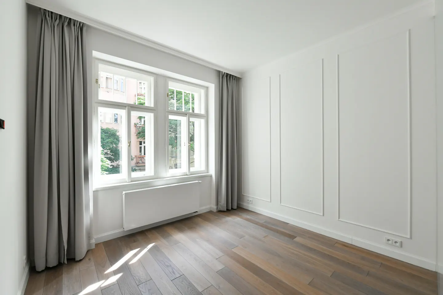 Bright, empty room with hardwood floors, white walls with molding, and a large window with gray curtains.