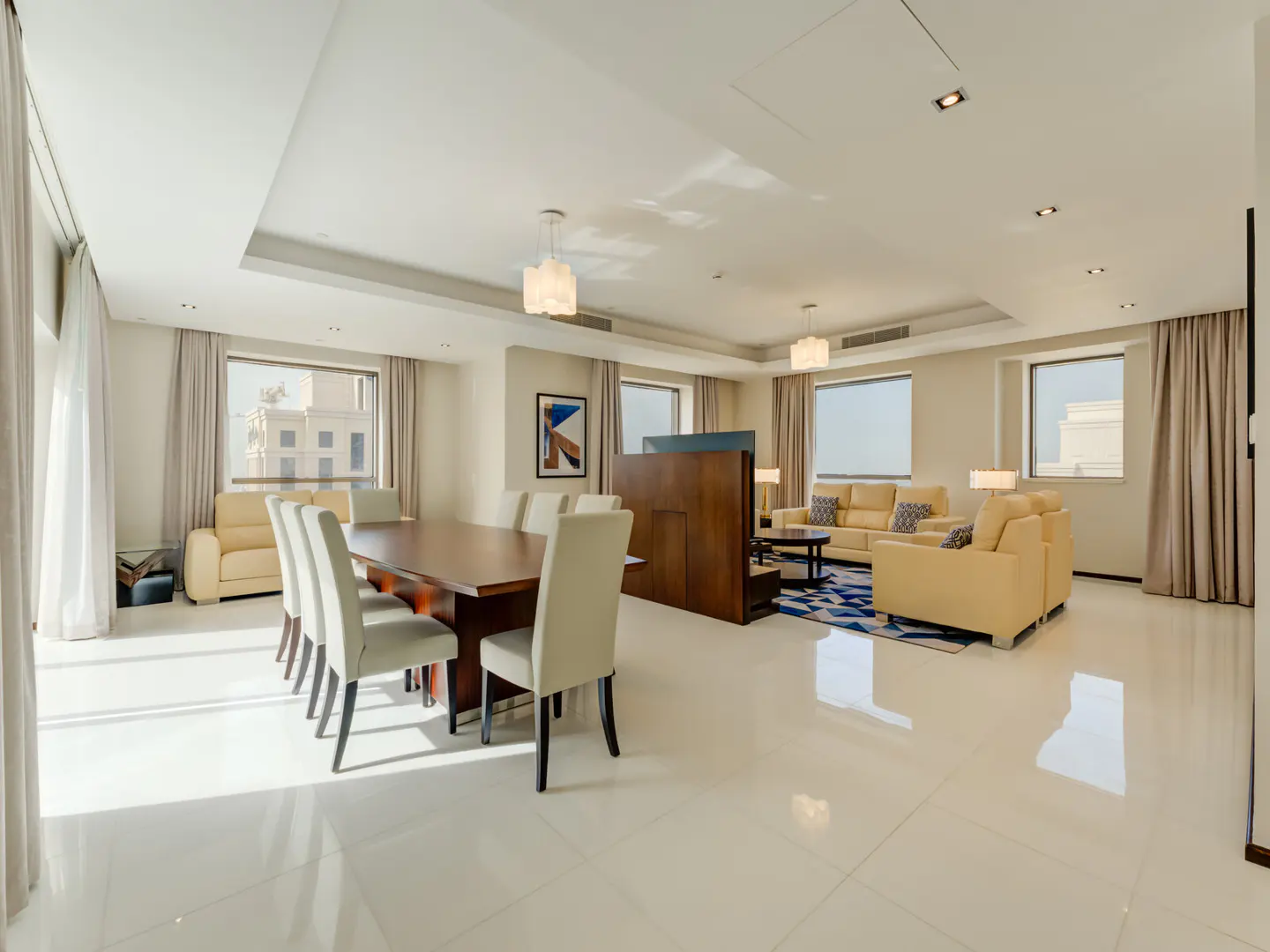 Bright, open-concept living and dining area with a wood table, cream chairs, and tan sofas on a blue patterned rug.
