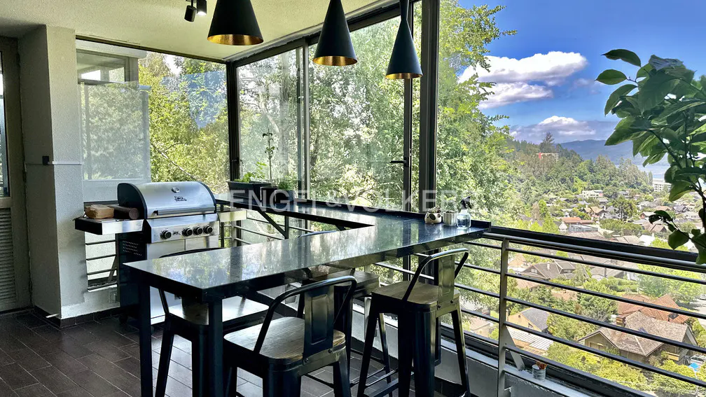 Indoor patio with grill, bar, and stools overlooking a lush green landscape and city view. Black pendant lights hang above.