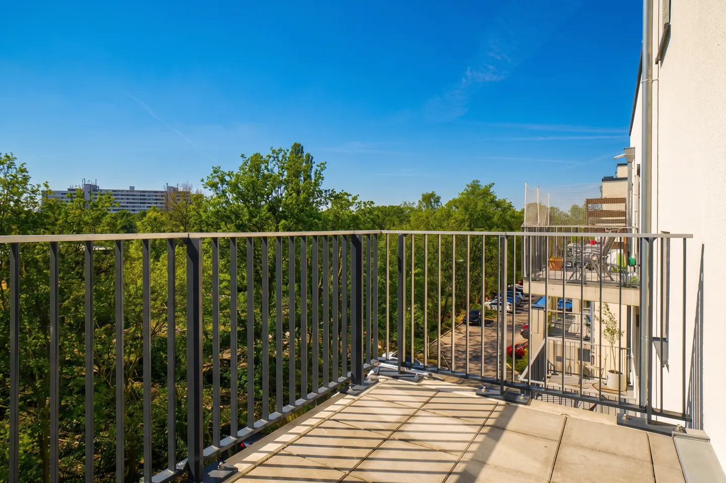 Balcony view with metal railings, tile floor, and shadows. Trees and a building are visible in the background under a blue sky.